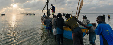 Several people push a boat out into a large body of water as the sun rises